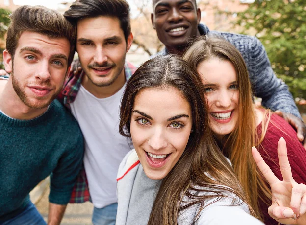 Cinq jeunes amis souriants prennent un selfie, l'une fait le signe de la paix.