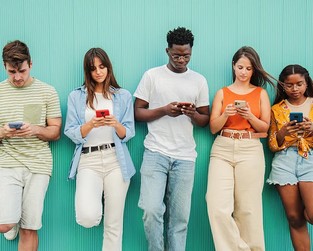 Cinq jeunes adultes de diverses origines utilisent leurs smartphones devant un mur vert.
