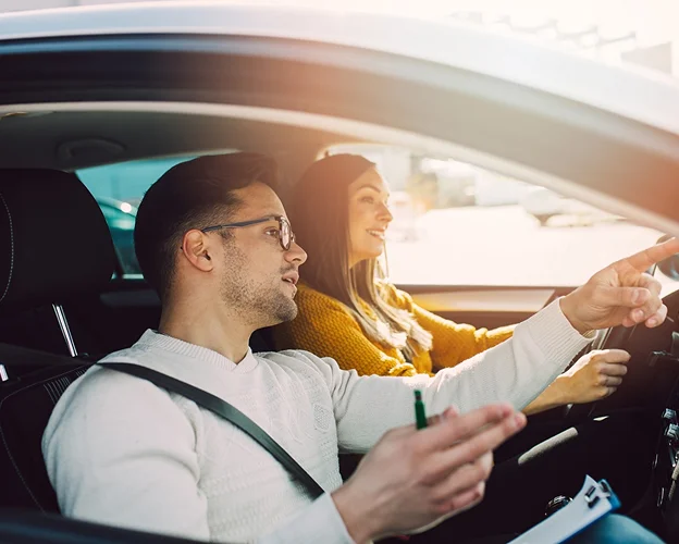 Moniteur d'auto-école pointant du doigt pendant qu'une élève conduit, souriante.