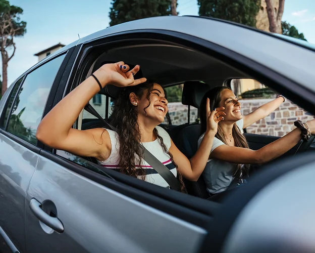 Deux jeunes femmes joyeuses conduisant une voiture, bras à l'extérieur, souriantes.