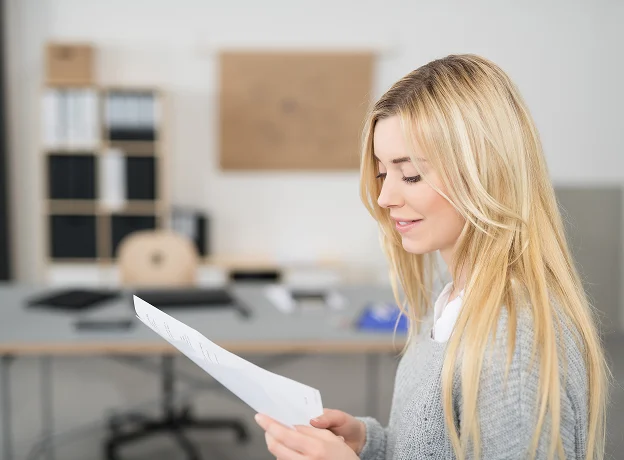 Jeune femme blonde souriante, les yeux baissés, lisant un document dans un bureau.